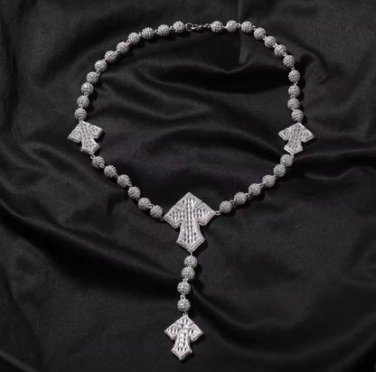 A silver-colored rosary bead chain necklace with baguette cut cubic zirconia stones and cross-shaped pendants, displayed on a black background.